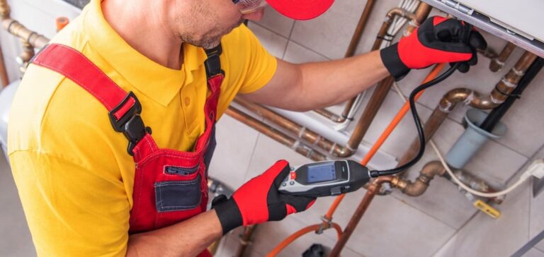 A professional plumber wearing red overalls and a yellow shirt uses an electronic leak detector to inspect copper pipes and a gas boiler system for repairs. professional plumbing services
